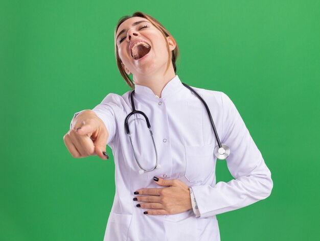 Lachende jonge vrouwelijke arts die medische mantel met een stethoscoop draagt, greep stoamch die je gebaar toont dat op groene muur wordt geïsoleerd
