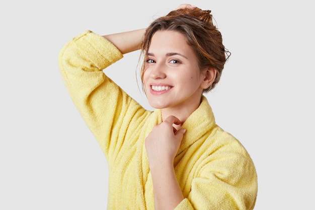 lachende meisje heeft geen make-up, gezonde huid, brede glimlach, gekleed in casual badjas, in een goed humeur na schoonheidsbehandelingen staat op witte studio muur. Gezichtsuitdrukkingen, beauty concept