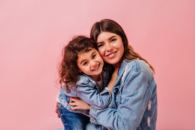 Lachende moeder en dochter camera kijken. Vooraanzicht van jonge vrouw met preteen kind dat op roze achtergrond wordt geïsoleerd.