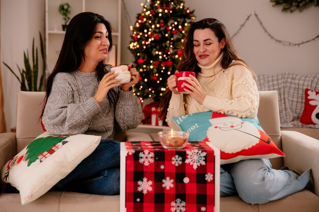 Lachende mooie jonge meisjes houden kopjes vast terwijl ze op fauteuils zitten en thuis genieten van kersttijd