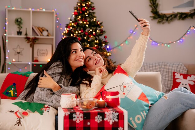 lachende mooie jonge meisjes nemen selfie zittend op fauteuils en genieten van kersttijd thuis