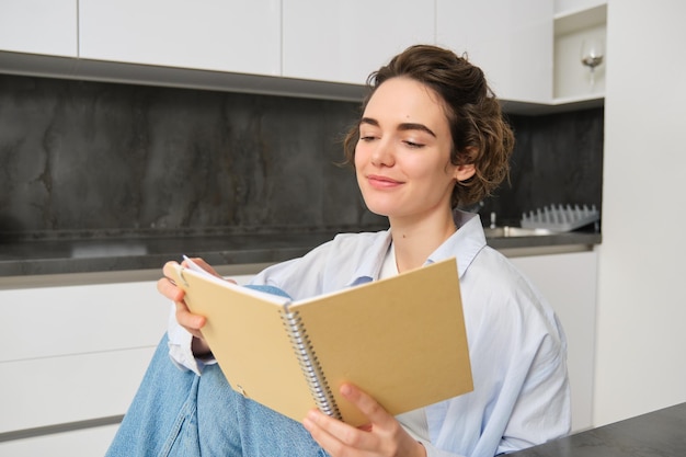 Lachende mooie vrouw zitten met notitieboekje in de keuken notities lezen studeren huiswerk