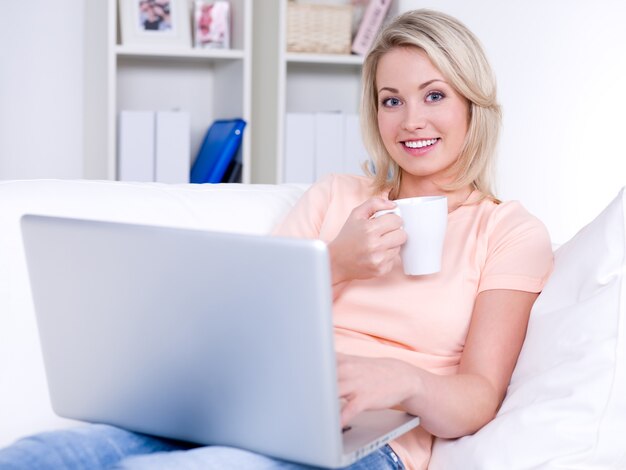 Lachende mooie vrouw zittend op de Bank met laptop en kopje koffie