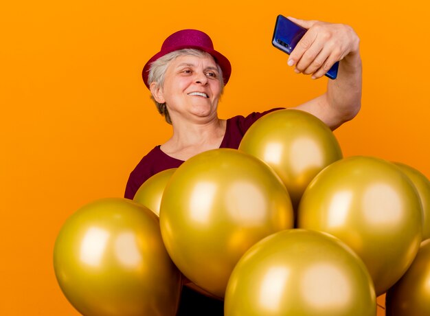 Gratis foto lachende oudere vrouw met feestmuts staat met helium ballonnen kijken naar telefoon geïsoleerd op oranje muur