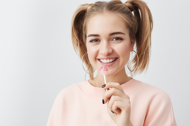 Lachende positieve vrouw met een aantrekkelijke uitstraling, het dragen van losse roze sweatshirt. Gelukkig tienermeisje met twee paardenstaarten op haar haren met positieve emoties eten lolly.