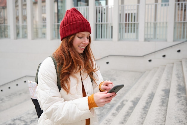 Gratis foto lachende roodharige studente toerist loopt door de stad, gaat de trap op en kijkt naar de kaart van de mobiele telefoon