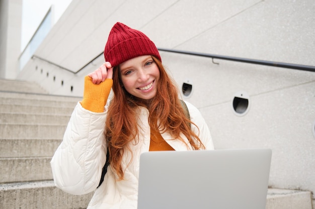 Lachende roodharige studente zit buiten op de trap en maakt gebruik van een laptop die verbinding maakt met openbare wifi in de stad