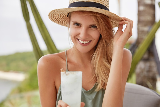 Lachende schattige vrouw met gelukkige uitdrukking heeft zomervakantie, besteedt vrije tijd op terras met vers koud drankje, ziet er positief uit. Aantrekkelijk wijfje dat in strohoed in goed humeur is.