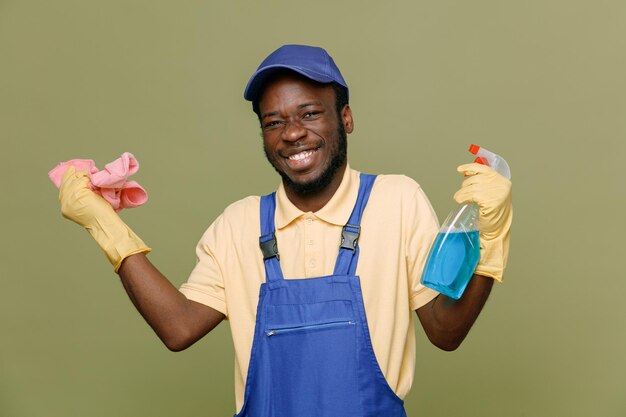 Lachende schoonmaakmiddel met vod jonge Afro-Amerikaanse schonere man in uniform met handschoenen geïsoleerd op groene achtergrond