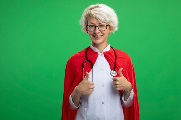 Lachende Slavische superheld vrouw in uniform van de arts met rode cape en stethoscoop in optische glazen duimen omhoog van twee handen geïsoleerd op groene muur met kopie ruimte