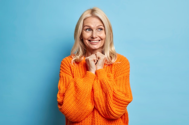 lachende tedere vrouw van middelbare leeftijd met licht haar brede glimlach houdt de handen bij elkaar en kijkt weg heeft een dromerige uitdrukking gekleed in een oversized trui staat over de blauwe muur.