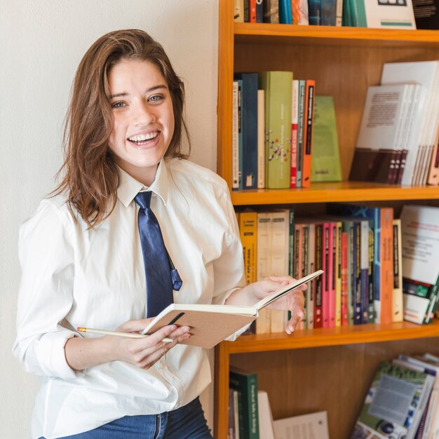 Lachende tiener met notitieboekje dichtbij boekenkast