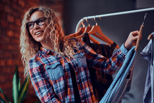 Lachende vrolijke vrouw in glazen en geruit hemd kiest kleding in butique voor haar vakantie. Ze heeft blond krullend haar. Vrouw kijkt weg.