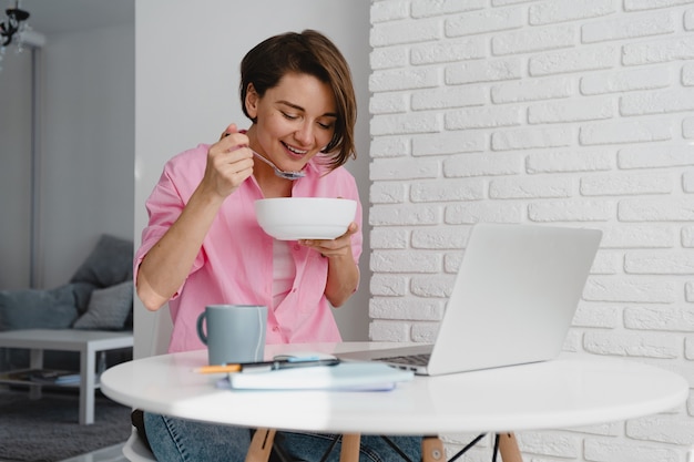 Lachende vrouw in roze shirt thuis ontbijten aan tafel online werken op laptop vanuit huis
