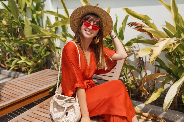 Lachende vrouw in stijlvolle oranje outfit en strooien hoed chillen op ligstoel in de buurt van zwembad.