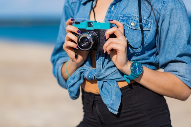 Lachende vrouw in strooien hoed en stijlvolle zomer outfit poseren met retro camera op het strand.