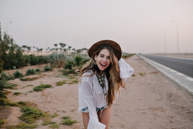 Lachende vrouw in trendy bruine hoed en witte blouse met plezier staande op zand naast snelweg. Portret van charmante langharige jonge vrouw buiten dansen en poseren met een glimlach, geniet van vakantie