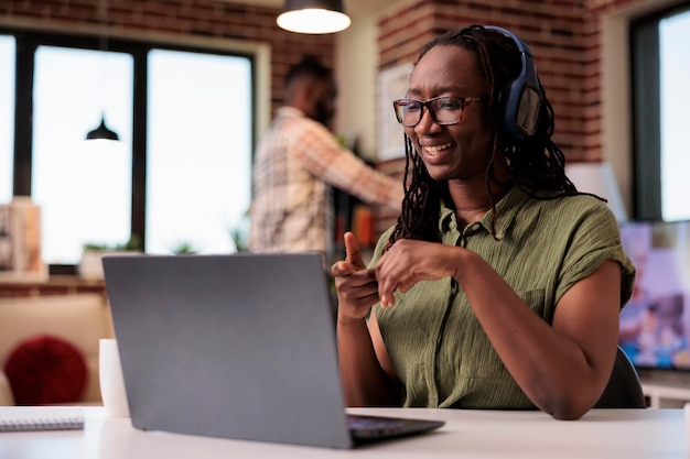 Lachende vrouw met draadloze koptelefoon kijken naar entertainmentshow op laptop terwijl ze aan een bureau zit. Afro-Amerikaanse freelancer die grappige sociale media-inhoud bekijkt op een draagbare computer.