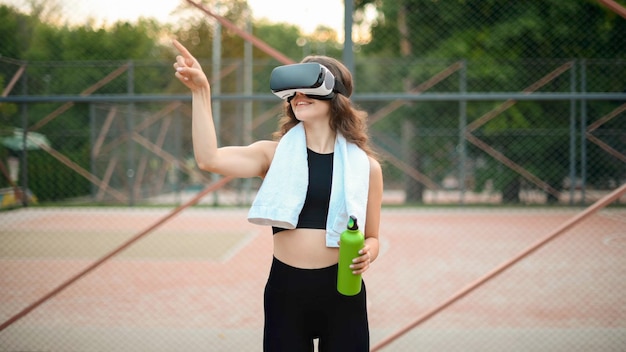 Gratis foto lachende vrouw met handdoek met vr-headset in een trainingspak met een waterfles op een sportveld