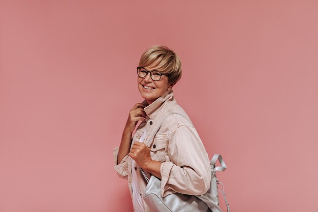 Lachende vrouw met stijlvolle blonde kapsel, bril en grijze tas in beige moderne jas op zoek naar camera op roze achtergrond.