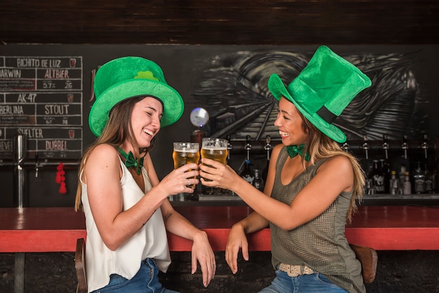 Lachende vrouwen in Saint Patricks hoeden die glazen drank klinken bij toog