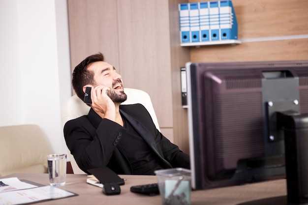 Lachende zakenman die aan de telefoon praat terwijl hij in zijn kantoor werkt. Ondernemer in professionele omgeving