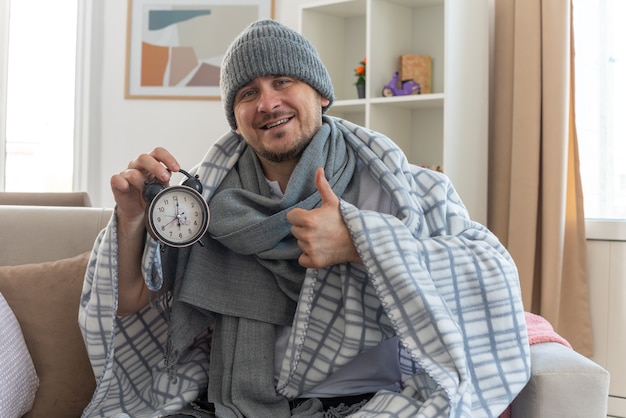 lachende zieke man met sjaal om nek met wintermuts gewikkeld in plaid met wekker en duimen omhoog zittend op de bank in de woonkamer