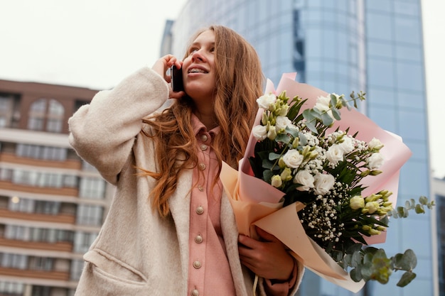 Lage hoek van elegante vrouw buitenshuis praten aan de telefoon en met een boeket bloemen