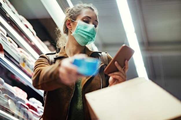 Lage hoekmening van een vrouw die een beschermend masker op haar gezicht draagt tijdens het gebruik van een mobiele telefoon en het kopen in de supermarkt tijdens een pandemie van het coronavirus