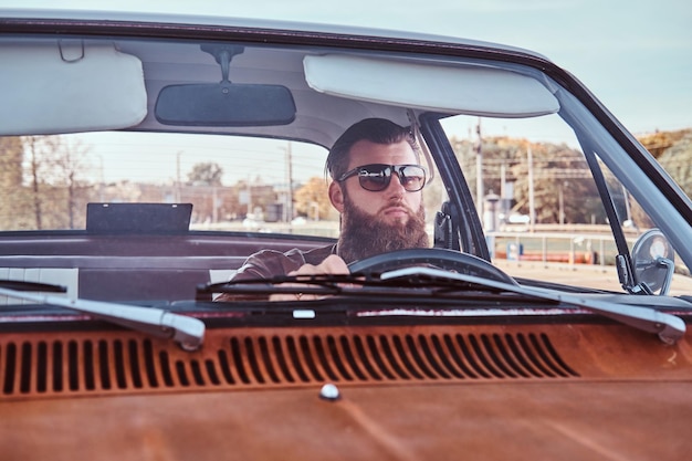 Landelijk stijlconcept. Bebaarde man in zonnebril gekleed in bruin lederen jas besturen van een retro auto.