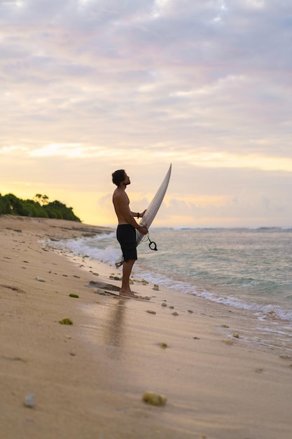 Landschapsbeeld van mannelijke surfer bezig met wandelen op het strand bij zonsopgang terwijl hij zijn surfplank onder zijn arm draagt met de oceaangolven die op de achtergrond breken. Jonge knappe mannelijke surfer op de oceaan