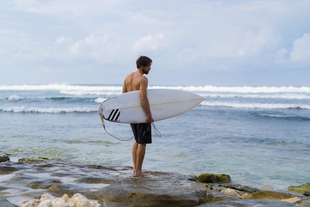 Gratis foto landschapsbeeld van mannelijke surfer bezig met wandelen op het strand bij zonsopgang terwijl hij zijn surfplank onder zijn arm draagt met de oceaangolven die op de achtergrond breken. jonge knappe mannelijke surfer op de oceaan
