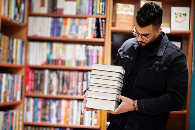 Lange slimme Arabische studentenman draagt een zwart spijkerjack en een bril in de bibliotheek met een stapel boeken