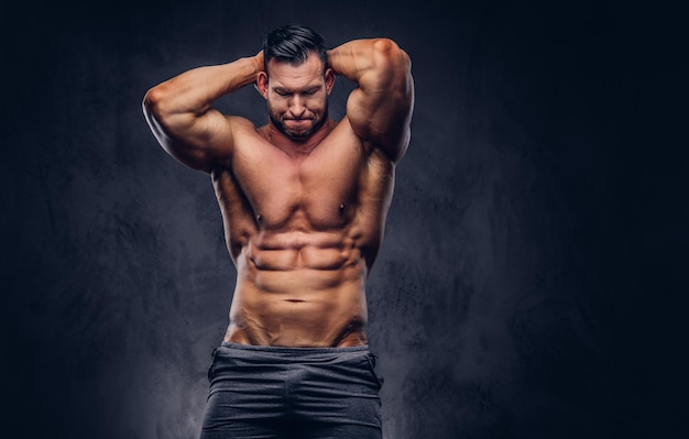 Lange stijlvolle shirtless bodybuilder gekleed in sportshorts, spieren tonend, staande in een studio. Geïsoleerd op een donkere achtergrond.