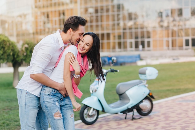 Langharig lachend meisje in gescheurde spijkerbroek met plezier met vriendje in stadspark. outdoor portret van grappige verliefde paar gek rond op straat met scooter op achtergrond.