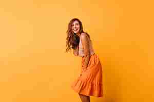 Gratis foto langharige krullende vrouw in lichte outfit die positieve emoties uitdrukt. romantisch vrouwelijk model in oranje rok die op geel danst.
