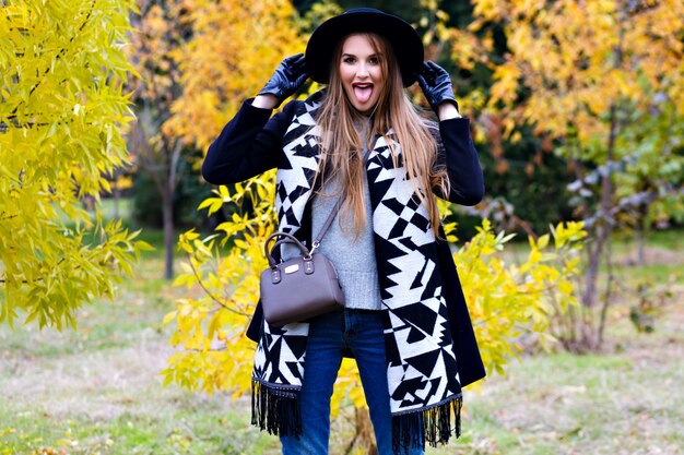 Langharige meisje draagt jeans spelen met haar sjaal in herfst park. Aantrekkelijke jonge vrouw in stijlvolle zwarte hoed tijd doorbrengen in bos en lachen.