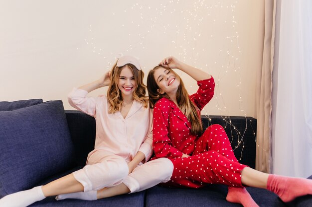Langharige meisje in roze sokken zittend op de bank met haar vriendin. Schattige jonge dames dragen nachtkostuums poseren op blauwe sofa.