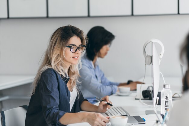 Langharige vrouwelijke programmeur met kopje koffie ontspannen van het werk. Indoor portret van Europese it-specialist zittend op de werkplek met glimlach naast Aziatische jongeman.