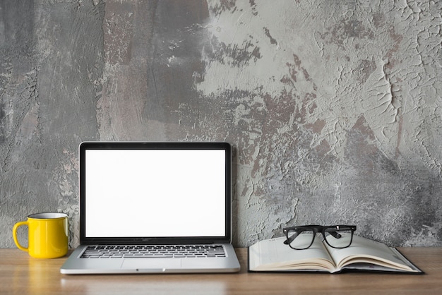 laptop; kop; boek en bril op houten bureau voor oude muur