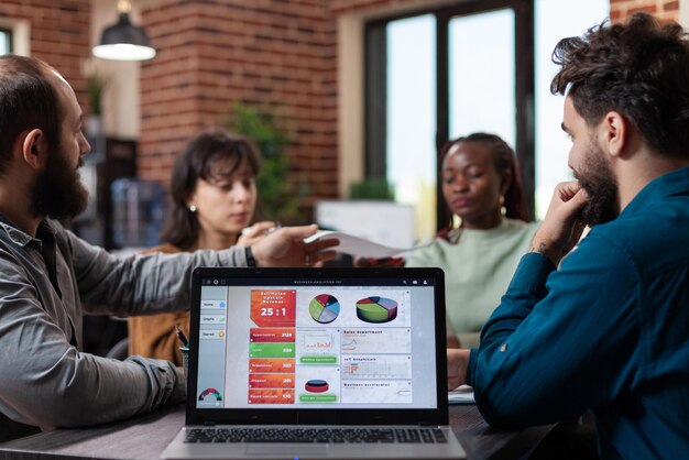 Laptopcomputer met marketinggrafieken op het scherm die op tafel in het opstartkantoor staan, terwijl diverse teams papieren delen met bedrijfsomzet erop. Multi-etnische zakenmensen die strategie analyseren