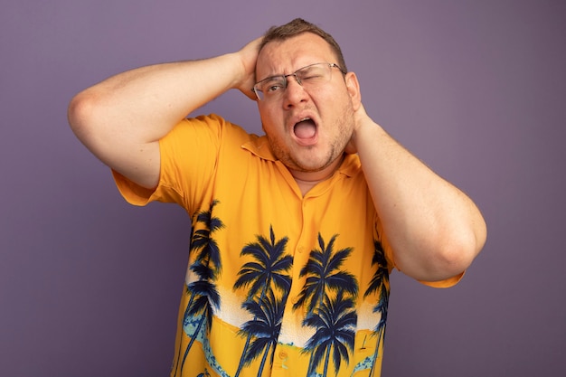 Lastige man in bril met oranje shirt op zoek verward en ontevreden zijn hoofd aan te raken dat over de paarse muur staat