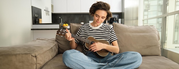 Leefstijl en hobby's concept jonge glimlachende vrouw op de bank die ukulele speelt zingt en leert