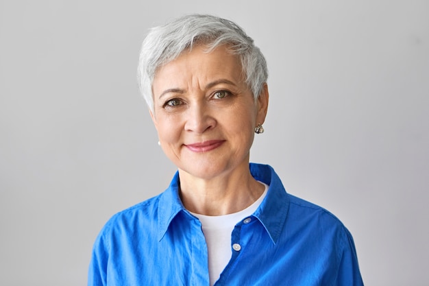 Leeftijd en schoonheidsconcept. Charmante positieve volwassen Europese vrouw met kort grijs haar en rimpels poseren geïsoleerde, zelfverzekerde glimlach, stijlvol blauw shirt dragen.