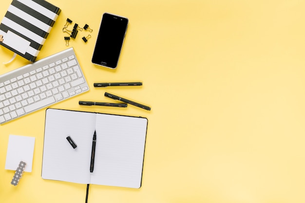 Leeg dagboek met pennen; bulldog paperclips; toetsenbord en mobiele telefoon op gele achtergrond