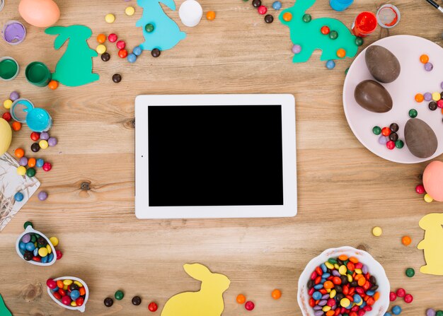 Lege digitale tablet omgeven met kleurrijke edelstenen snoepjes; Paas eieren; papieren knipsel konijn op houten tafel