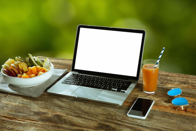 Lege schermen van laptop en smartphone op een houten tafel buiten met natuur op muur Fruit en vers sap in de buurt. Concept van creatieve werkplek, zaken, freelance. Copyspace.