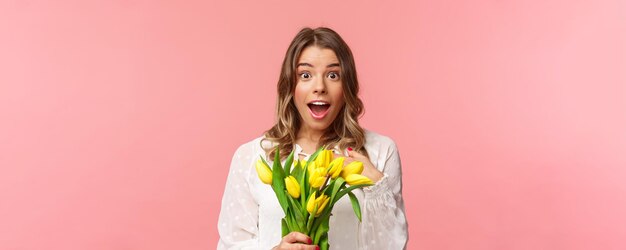 Lente geluk en viering concept Close-up van verrast en verbaasd jong blond meisje in witte jurk ontvangt gele tulpen en wijst zichzelf met ongeloof en verbazing had niet verwacht