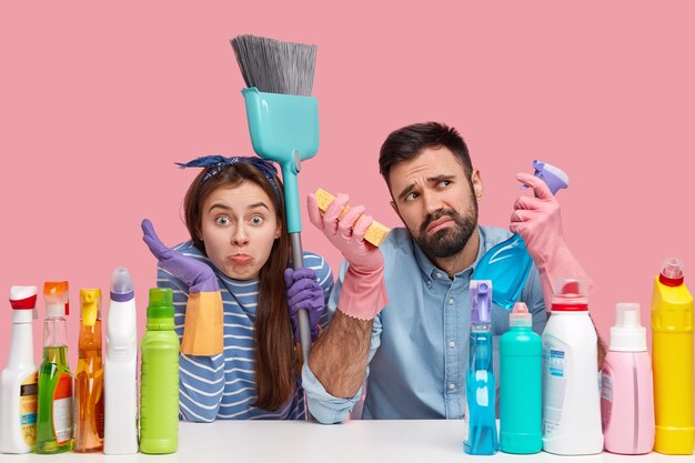 Lente schoonmaak concept. Horizontale opname van man en vrouw houden spray wasmiddel, omgeven met schoonmaakbenodigdheden, bezem gebruiken, draagt rubberen handschoenen voor handbescherming, huis samen opruimen