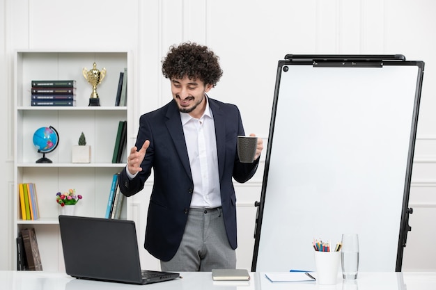 Leraar brunette instructeur met computer in pak en whiteboard in klaslokaal met koffiekopje